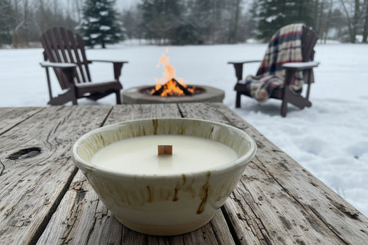 Candle in a white bowl on a wooden table with a snowy landscape and fire pit in the background.
